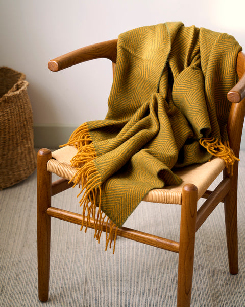 Mustard colour knitted blanket draped over a wooden chair with a woven basket in the background.