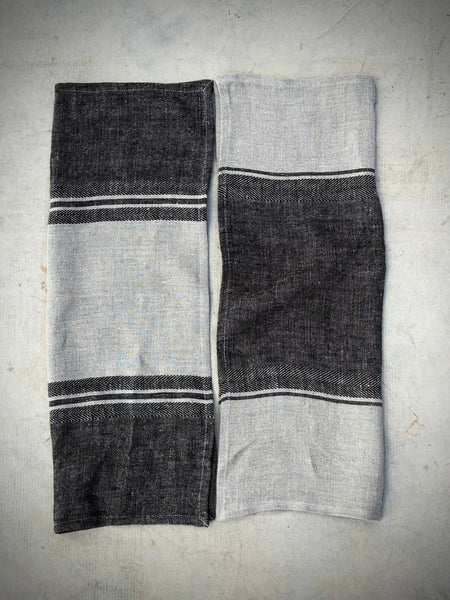 Two folded linen napkins with a black and light gray herringbone pattern and thin white stripe accents, displayed side-by-side on a textured gray surface.