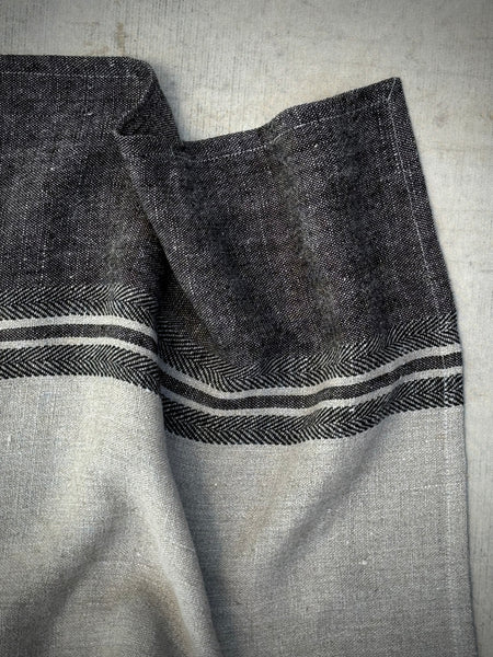 A close-up of a woven textile, likely a table runner or napkin, featuring a dark charcoal gray herringbone pattern with two horizontal white stripes, set against a light gray linen background.