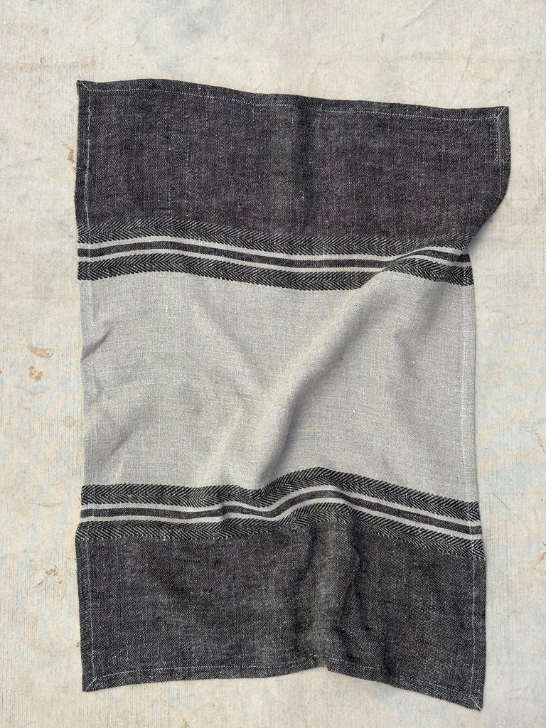 A rectangular woven textile towel  laid flat on a light textured surface. The design features a large light gray center panel bordered by dark charcoal gray sections at the top and bottom, with thin white stripe accents near the borders.