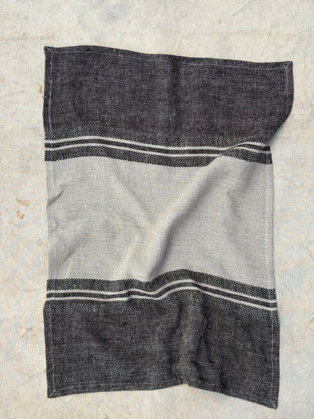 A rectangular woven textile towel  laid flat on a light textured surface. The design features a large light gray center panel bordered by dark charcoal gray sections at the top and bottom, with thin white stripe accents near the borders.